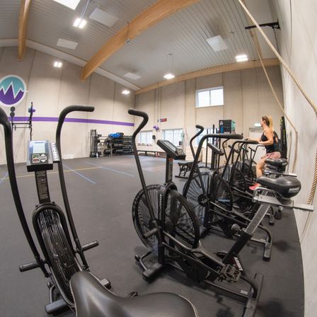 Ein Fitnessstudio mit verschiedenen Trainingsgeräten, darunter Laufbänder und Rudergeräte, hat eine Frau, die auf einem Rudergerät sitzt. Das Fitnessstudio hat Holzbalken, weiße Wände und Lichter an der Decke.