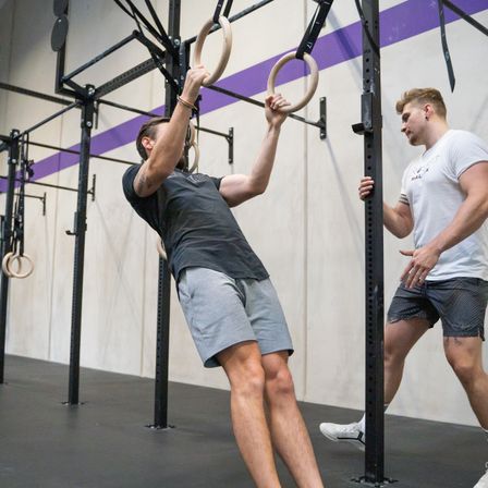 Zwei Männer trainieren in einem Fitnessstudio. Einer zieht sich an einem Ring an einer Stange hoch, während der andere zuschaut.