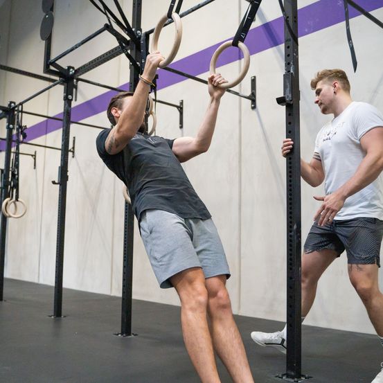 Zwei Männer trainieren in einem Fitnessstudio. Einer zieht sich an einem Ring an einer Stange hoch, während der andere zuschaut.