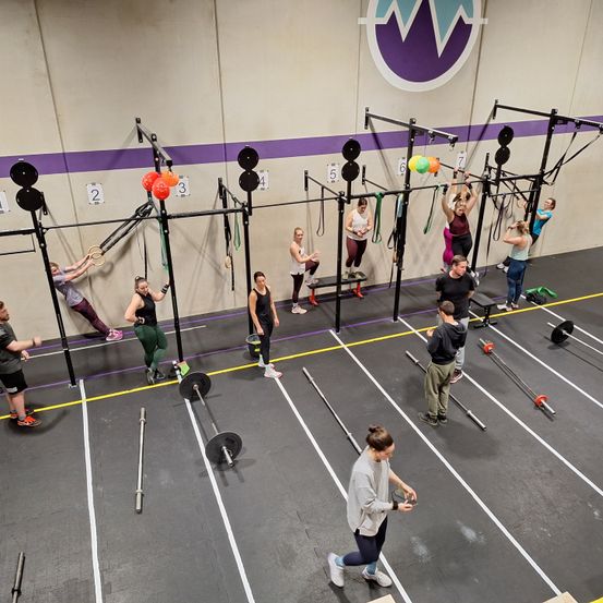 Eine Gruppe von Menschen in einem Fitnessstudio macht verschiedene Übungen mit einer Hantel und einem Trapez. Einige tragen Turnschuhe, während andere keine Schuhe tragen.