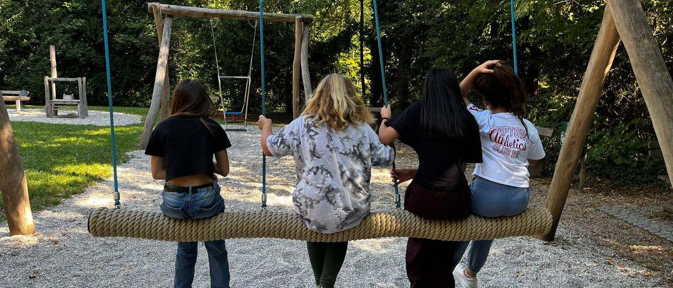 Vier Mädchen sitzen auf einer Seilschaukel in einem Park. Zwei von ihnen haben die Hände auf dem Kopf. Hinter ihnen ist eine weitere Schaukel zu sehen. Bäume und Gras umgeben das Gebiet.