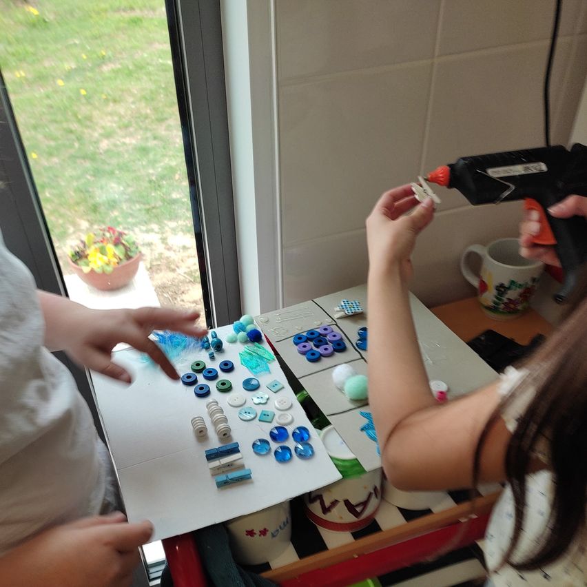 Two kids use a hot glue gun to glue beads on a paper sheet in a room with a window showing a lawn.