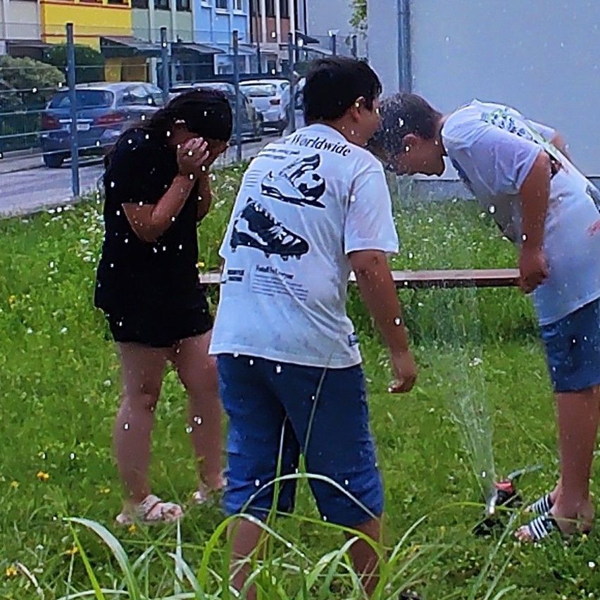 Drei junge Leute spielen auf einer Rasenfläche mit einem Wasserstrahl. Ein Junge lehnt sich vorwärts, während ein anderer zurückgelehnt steht. Ein Mädchen steht in der Nähe und bedeckt ihr Gesicht. Autos sind am Straßenrand geparkt.