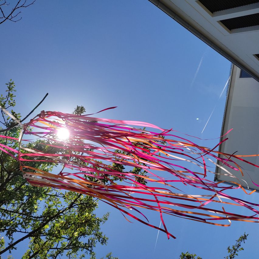 Ein farbenfrohes, buntes Bänder-Dekoration hängt von einem Ast unter einem klaren blauen Himmel. Die Sonne scheint hell und beleuchtet die Bänder.