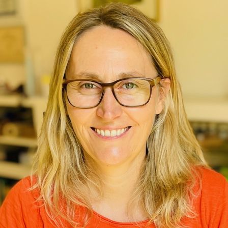 Eine Frau mit blonden Haaren und einer Brille lächelt in einer Innenumgebung. Sie trägt ein orangefarbenes Shirt. Hinter ihr ist ein verschwommener Hintergrund mit Regalen und Gegenständen.