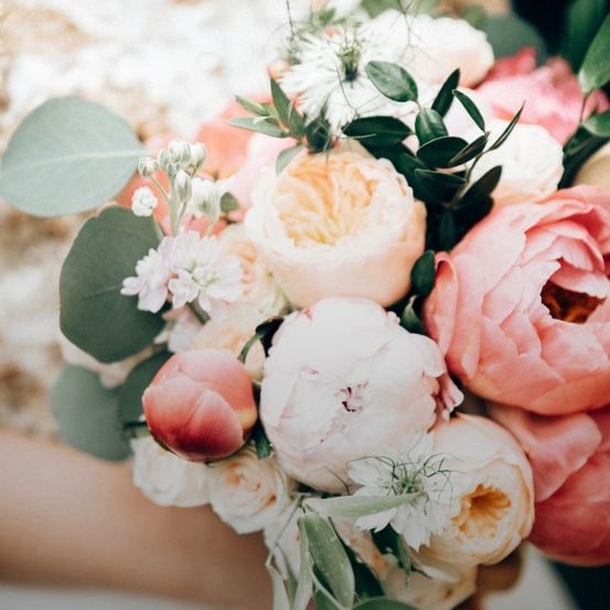 Eine Nahaufnahme eines Hochzeitsbouquets mit rosa, weißen und pfirsichfarbenen Rosen und grünen Blättern. Das Bouquet wird von einer Person gehalten.