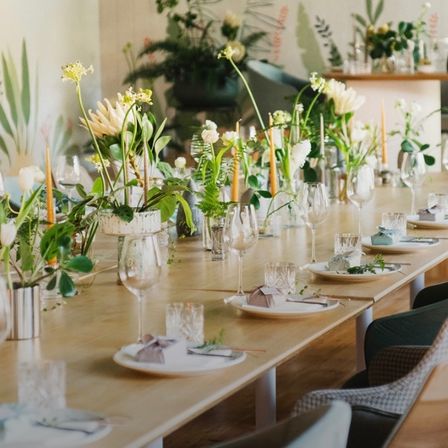 Ein langer Esstisch, der für eine Party gedeckt ist, mit weißen Tellern, Weingläsern und verschiedenen Blumenarrangements. Kerzen und Grünpflanzen schmücken den Tisch, während Pflanzen und dekorative Elemente im Hintergrund stehen.