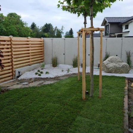 Ein Garten mit einem Holzzaun, einem großen Felsen und einem Baum, der von Holzpfosten gestützt wird. Der Rasen ist gut gepflegt, und es gibt Pflanzen und Bäume im Hintergrund.