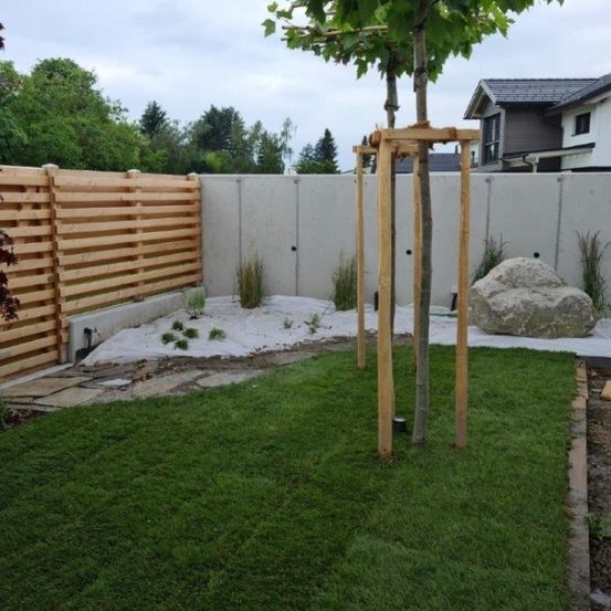 Ein Garten mit einem Holzzaun, einem großen Felsen und einem Baum, der von Holzpfosten gestützt wird. Der Rasen ist gut gepflegt, und es gibt Pflanzen und Bäume im Hintergrund.