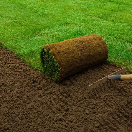 Ein gerollter Grasrasen liegt neben einem grünen Rasen auf der Erde, mit einem Rechen auf dem Boden.