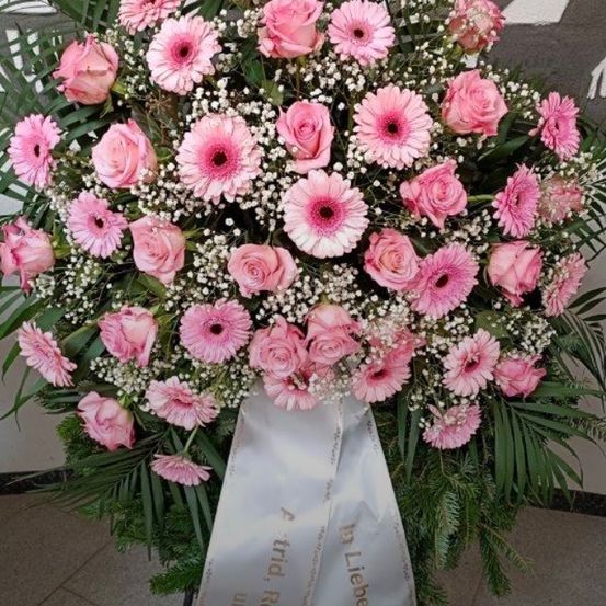 Ein großer Strauß aus rosa Rosen und Gänseblümchen mit einer weißen Schleife, die 'In lieber Erinnerung' steht. Der Strauß ist mit Babysate und grünen Blättern arrangiert.