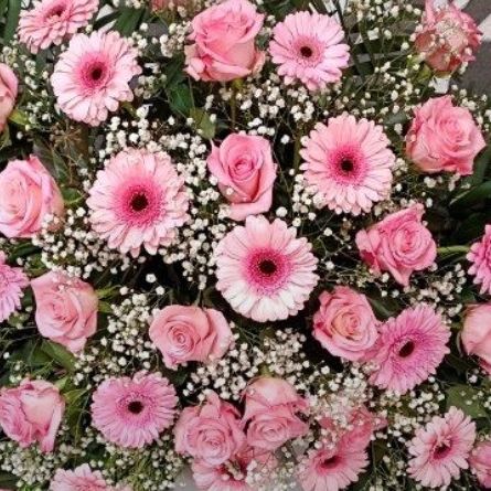 Eine Blumenarrangement mit rosa Rosen und Gänseblümchen, verziert mit Babysatem und grünen Blättern, auf einem grauen Hintergrund ausgestellt. Eine weiße Schleife mit dem Text 'In Liebe' ist an den Blumen befestigt.