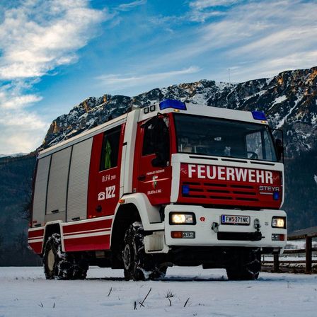 A red and white fire truck with snow tires is parked in a snowy area with a fence and mountains in the background.
