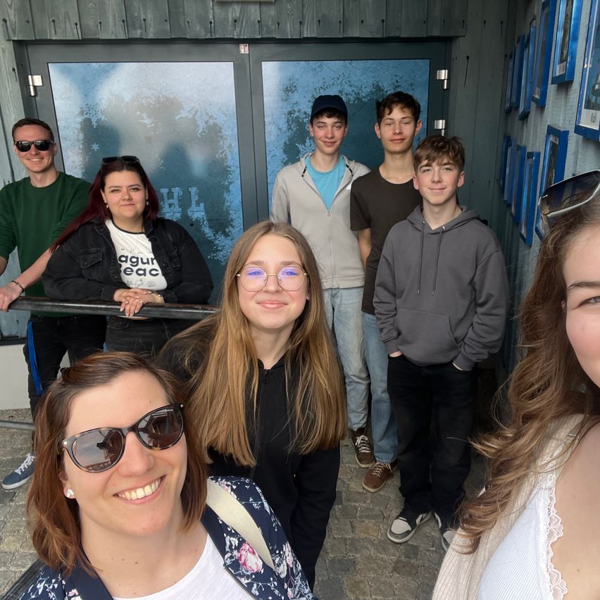 A group of people are standing in front of a building, smiling and posing for a picture. Three men are standing behind a woman with glasses, and they are all looking at the camera.