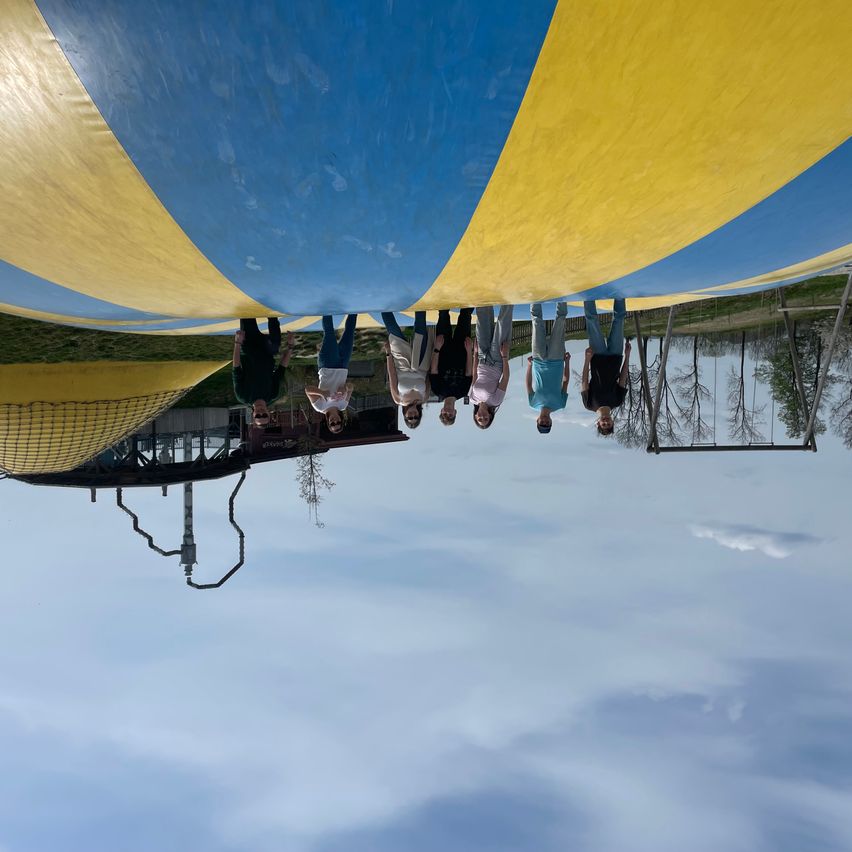 A group of people stand beneath a blue and yellow inflatable slide, looking upwards. The sky is cloudy.