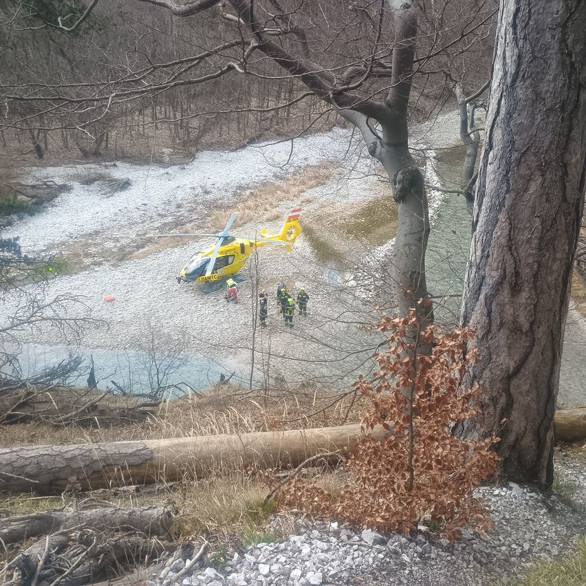 Ein gelber Hubschrauber ist am Flussufer geparkt, in der Nähe steht eine kleine Gruppe von Personen in Schutzausrüstung.