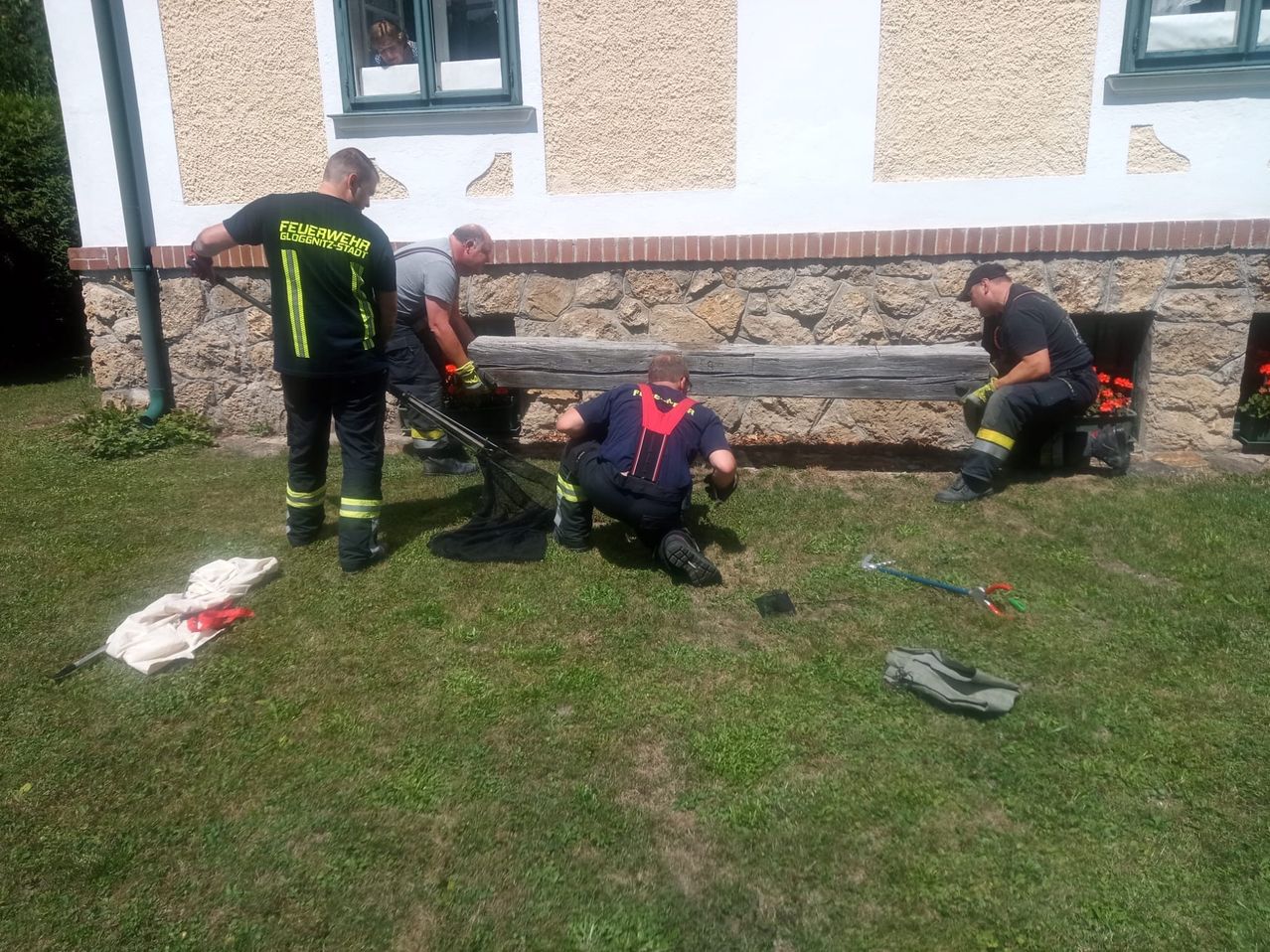 Vier Feuerwehrleute arbeiten an einer Holzstruktur auf einem Rasen. Einer kniet und hält ein Werkzeug, während die anderen stehen und Werkzeuge halten. Eine Person beobachtet aus einem Fenster.
