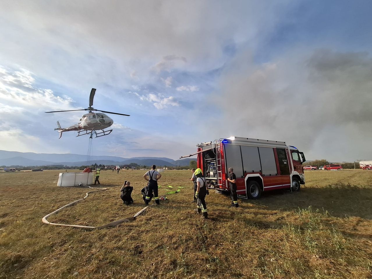 Ein Löschhubschrauber schwebt über einem Feld, während Feuerwehrleute am Boden stehen, einer hält eine Feuerwehrschlauch. Ein Feuerwehrwagen ist geparkt, und Rauch steigt im Hintergrund auf.