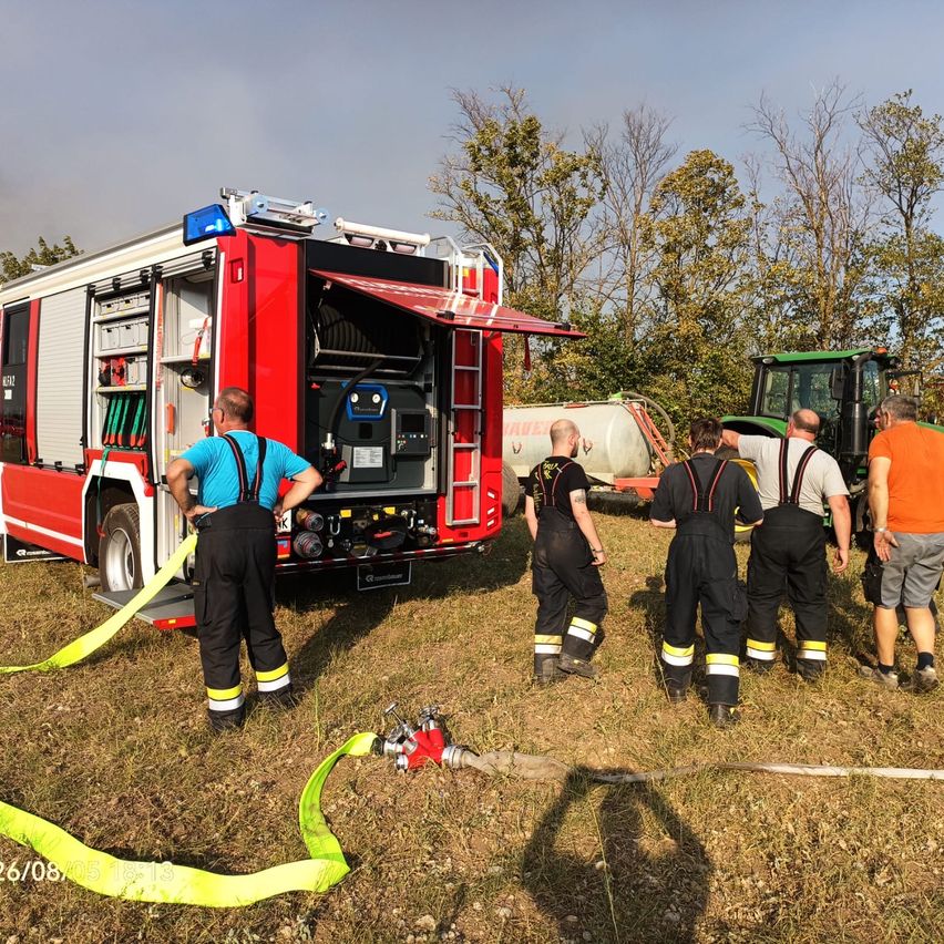 Eine Gruppe von Feuerwehrleuten in schwarzen Uniformen steht um einen roten Feuerwehrwagen mit offener Tür herum, während ein Traktor in der Nähe geparkt ist.