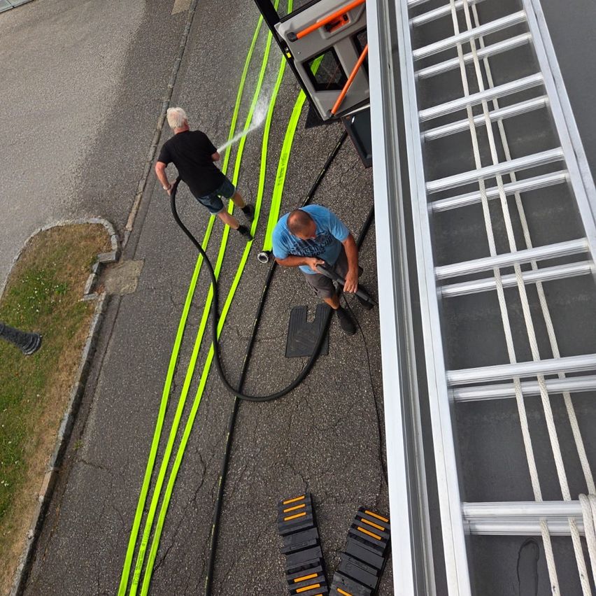 Two men are working on the side of a building, one spraying water from a hose and the other holding a tool. The scene is set on a road with yellow and green markings.
