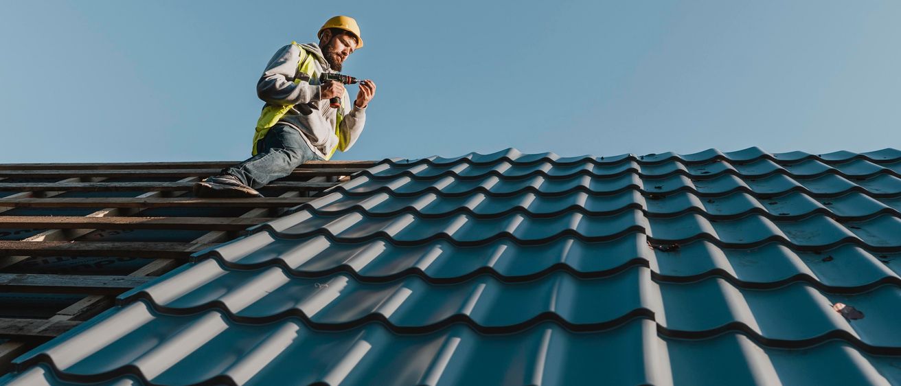 Ein Mann auf einem Dach, trägt einen gelben Schutzhelm, hält eine Bohrmaschine und arbeitet unter einem blauen Himmel.
