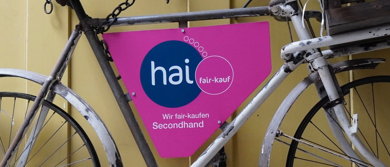 Ein altes Fahrrad mit einem rosa Schild, das 'hai fair-kauf Secondhand' sagt, hängt an einer gelben Wand. Das Fahrrad hat einen braunen Sattel, rostige Ketten und einen Holzkorb.
