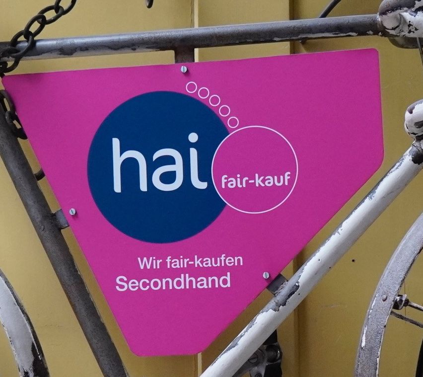Ein altes Fahrrad mit einem rosa Schild, das 'hai fair-kauf Secondhand' sagt, hängt an einer gelben Wand. Das Fahrrad hat einen braunen Sattel, rostige Ketten und einen Holzkorb.