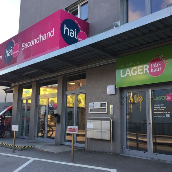 Der Eingang zu einem Secondhand-Laden namens 'hai' ist zu sehen. Das Schild ist pink mit dem Wort 'Secondhand'. Ein grünes Schild mit dem Wort 'Lager' ist daneben. Der Laden hat Glastüren und Fenster. Fahrräder sind draußen geparkt.