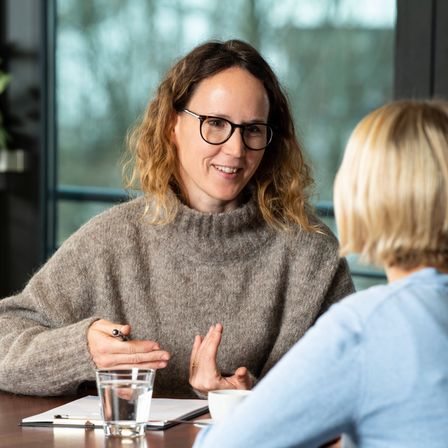 Eine Frau mit Brille lächelt, während sie einen Stift hält und mit einer anderen Frau am Tisch spricht, auf dem ein Glas Wasser und ein Notizbuch stehen.