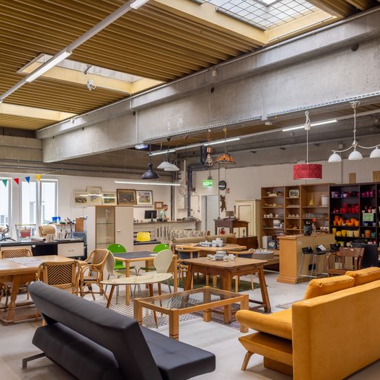 Ein gut beleuchteter Indoor-Möbelausstellungsraum mit Holztischen, Stühlen und Sofas. Der Raum hat mehrere Lampen, die von der Decke hängen, und verschiedene Gegenstände auf Regalen.