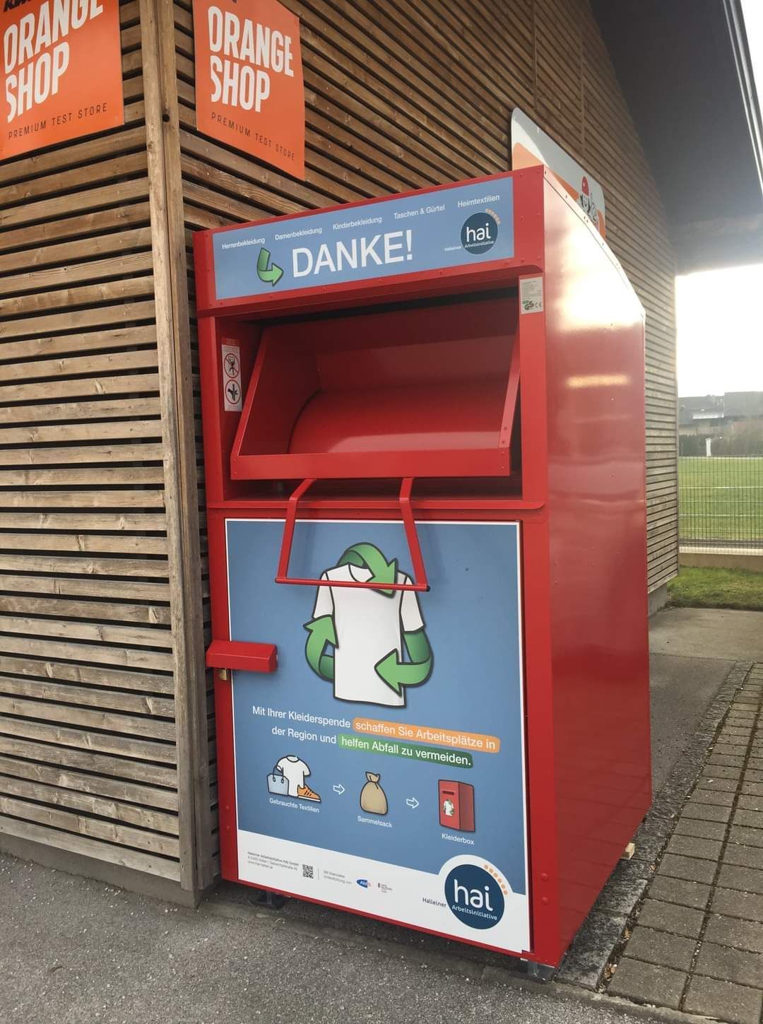 Ein roter Spendenautomat für Kleidung steht an der Seite eines hölzernen Gebäudes. Er hat ein blaues Schild mit Danke! und einem grünen Recycling-Symbol.