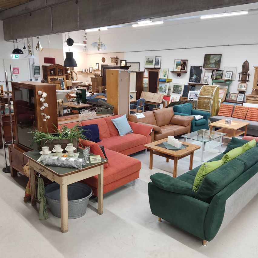 Ein Raum mit verschiedenen Möbeln, darunter Sofas, Tische und Dekorationsartikel, ist gut beleuchtet mit Deckenleuchten. Der Raum hat eine Mischung aus modernen und Vintage-Möbeln.