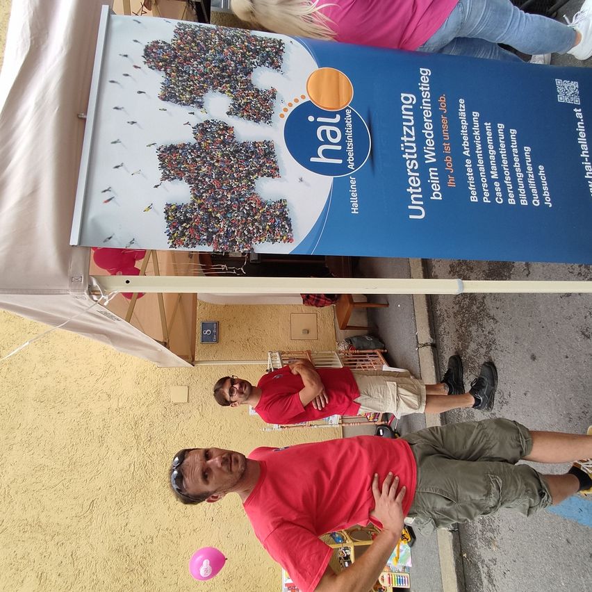 Zwei Männer in roten Shirts und khakifarbenen Shorts posieren für ein Foto unter einem Banner, das eine Jobmesse mit einer Menschenmenge-Illustration bewirbt.