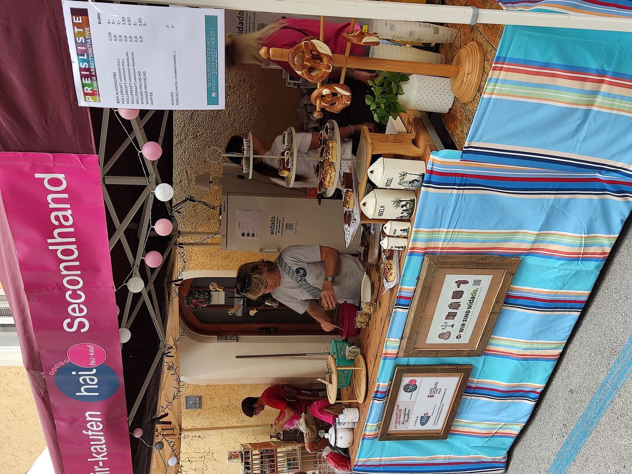 Ein Bäckereistand wird von oben gezeigt, auf dem verschiedene Backwaren auf Tischen ausgestellt sind. Kunden sind sichtbar, einer greift nach einem Gebäck, während ein anderer sitzt. Der Hintergrund umfasst eine gestreifte Tischdecke und ein Banner mit der Aufschrift 'Fair-kaufen'.