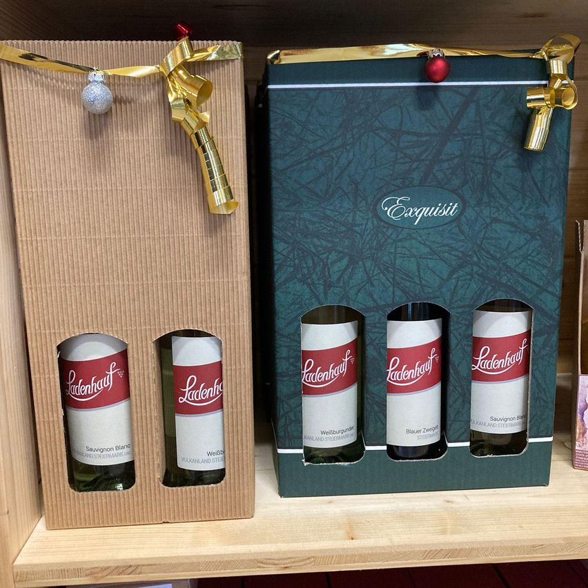 Zwei Weingeschenksets auf einem Holzregal. Eine Box enthält drei Ladenhof Weißweinflaschen, die andere vier mit roten Bändern. Beide haben dekorative Ornamente.