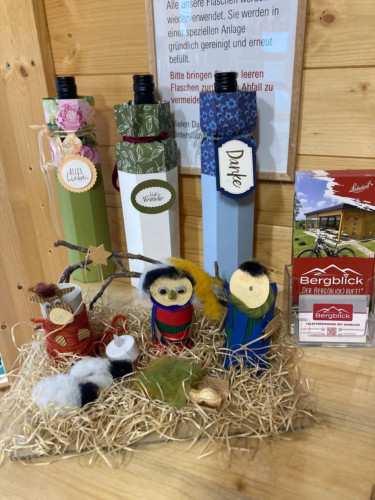 Drei dekorierte Weinflaschen stehen auf einer Holzoberfläche. Eine Weihnachtsszene aus Wolle und Holz befindet sich in der Mitte, mit einem kleinen Baum und einem Stern. Eine rote Box ist rechts.