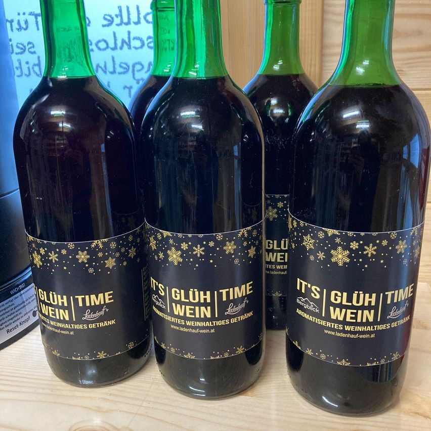 Fünf Flaschen aromatischer Glühwein mit dekorativen Schneeflocken auf schwarzen Etiketten sind auf einer Holzoberfläche aufgereiht.