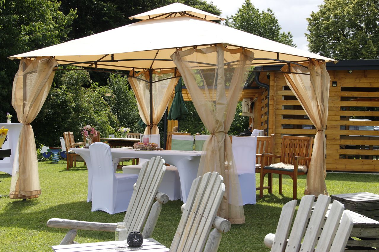 Ein Outdoor-Gazebo mit weißen Stühlen und Tisch unter einem großen weißen Baldachin. Es gibt zwei weiße Stühle vorne und eine schwarze Kerze auf dem Tisch.