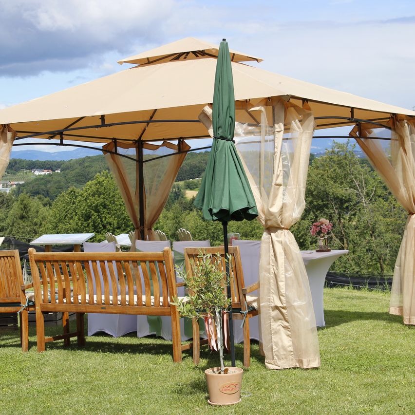 Ein Gartenpavillon mit Tisch und Stühlen. Der Pavillon ist mit beigem Stoff bedeckt und hat einen grünen Schirm. Ein Topf mit Pflanzen steht vor dem Pavillon auf dem Rasen.