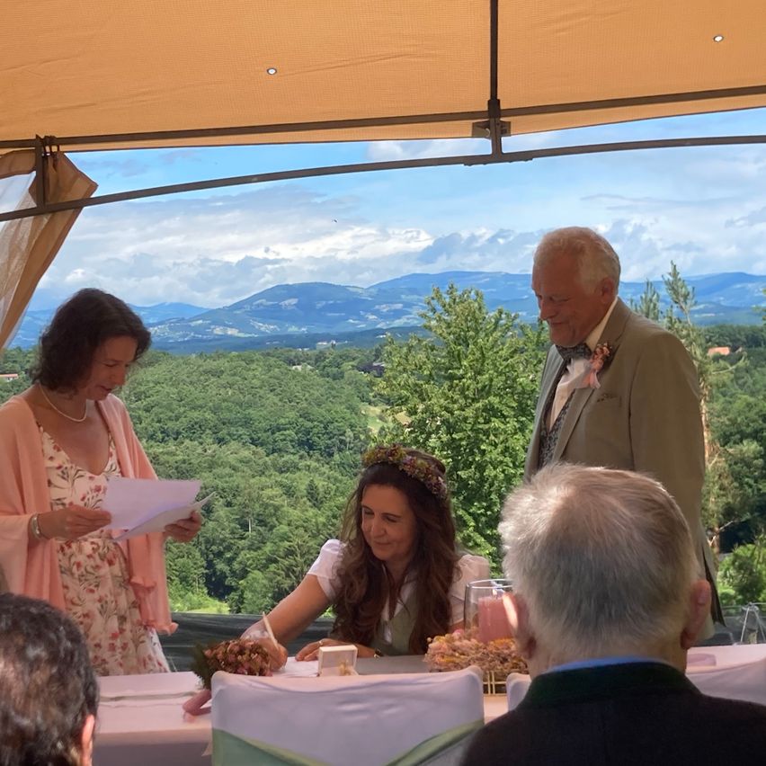 Eine Hochzeitszeremonie im Freien mit einer Frau, die aus einem Papier liest, einer Frau mit einer Blumenkopfbedeckung beim Unterschreiben und zwei älteren Männern. Sie stehen unter einem Zelt mit Blick auf Berge.