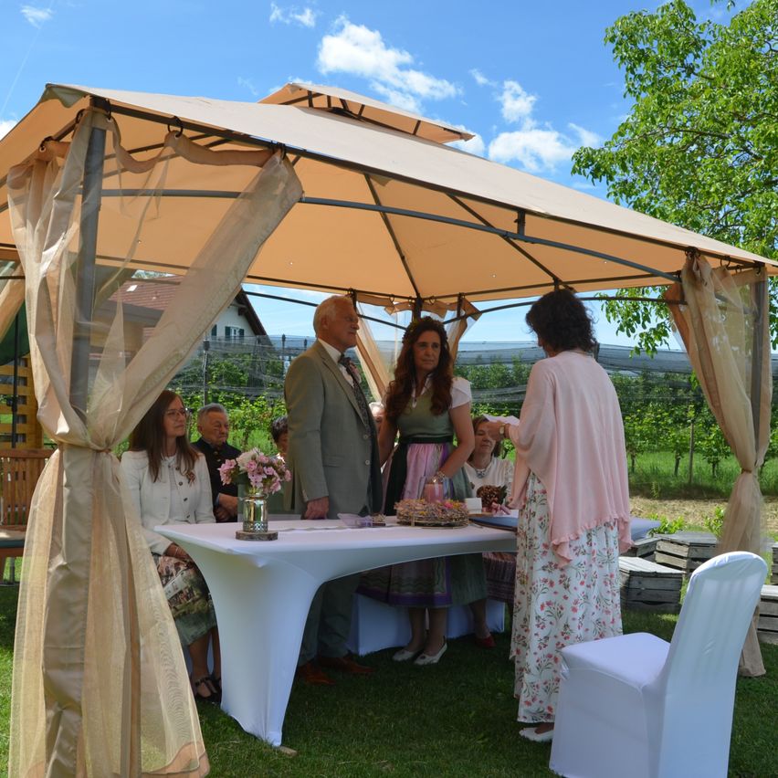 Eine Hochzeitszeremonie unter einem Baldachin mit Blumendekorationen und sitzenden Gästen. Die Braut steht neben einem Tisch mit Blumen, und der Bräutigam steht mit dem Rücken zur Kamera.