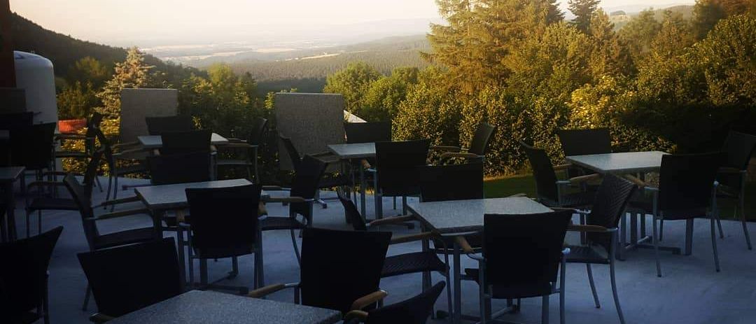 Eine Terrasse mit mehreren leeren Tischen und Stühlen, umgeben von Bäumen und einem entfernten Tal. Der Himmel ist bedeckt.