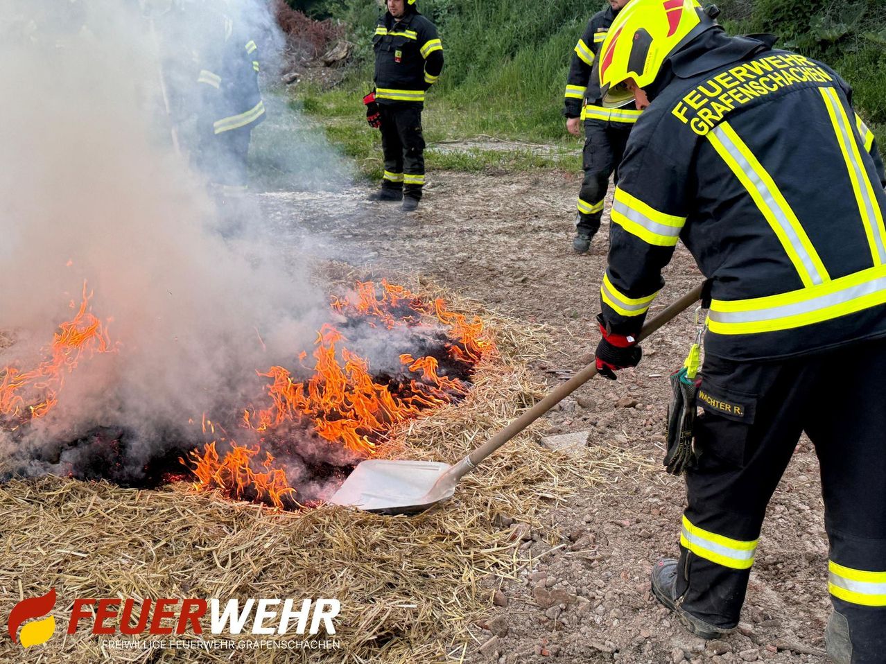 Ein Feuerwehrmann mit gelbem Helm löscht ein Feuer mit einer Schaufel auf einem Feld. Andere Feuerwehrleute stehen hinter ihm und beobachten.