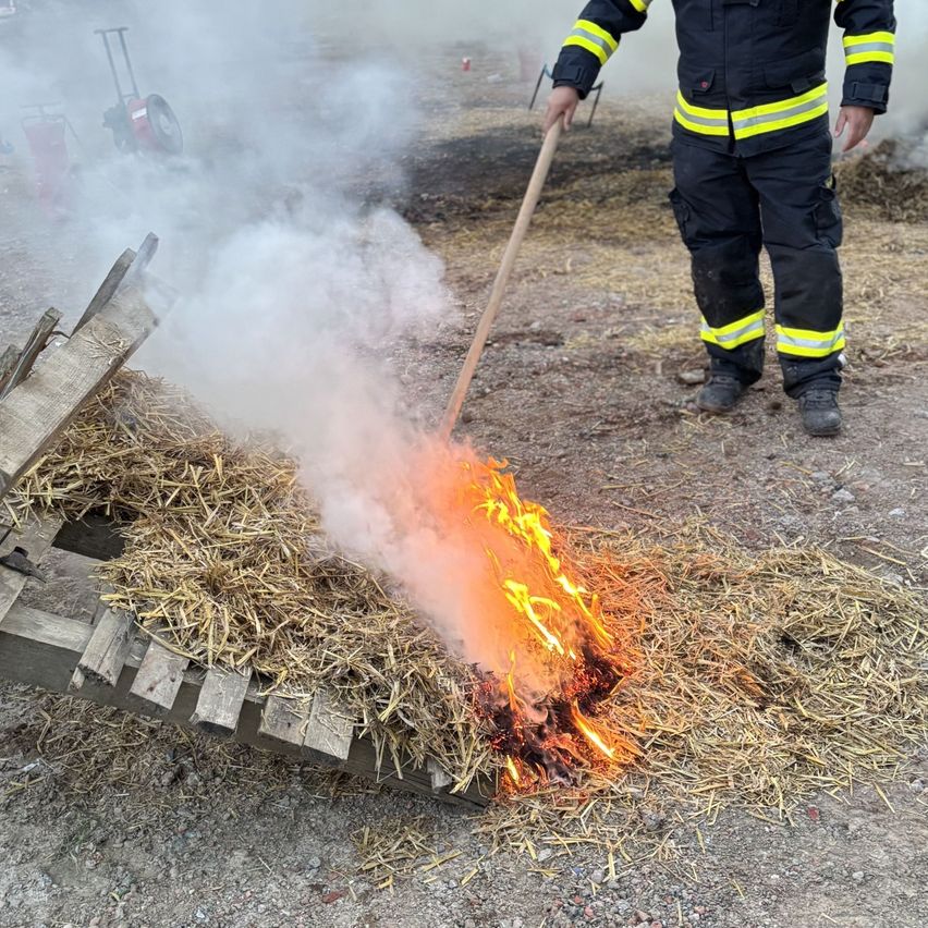 Ein Feuerwehrmann benutzt einen Stock, um ein kleines Feuer in einem Feld mit trockenem Gras zu löschen. Rauch steigt aus den Flammen auf.