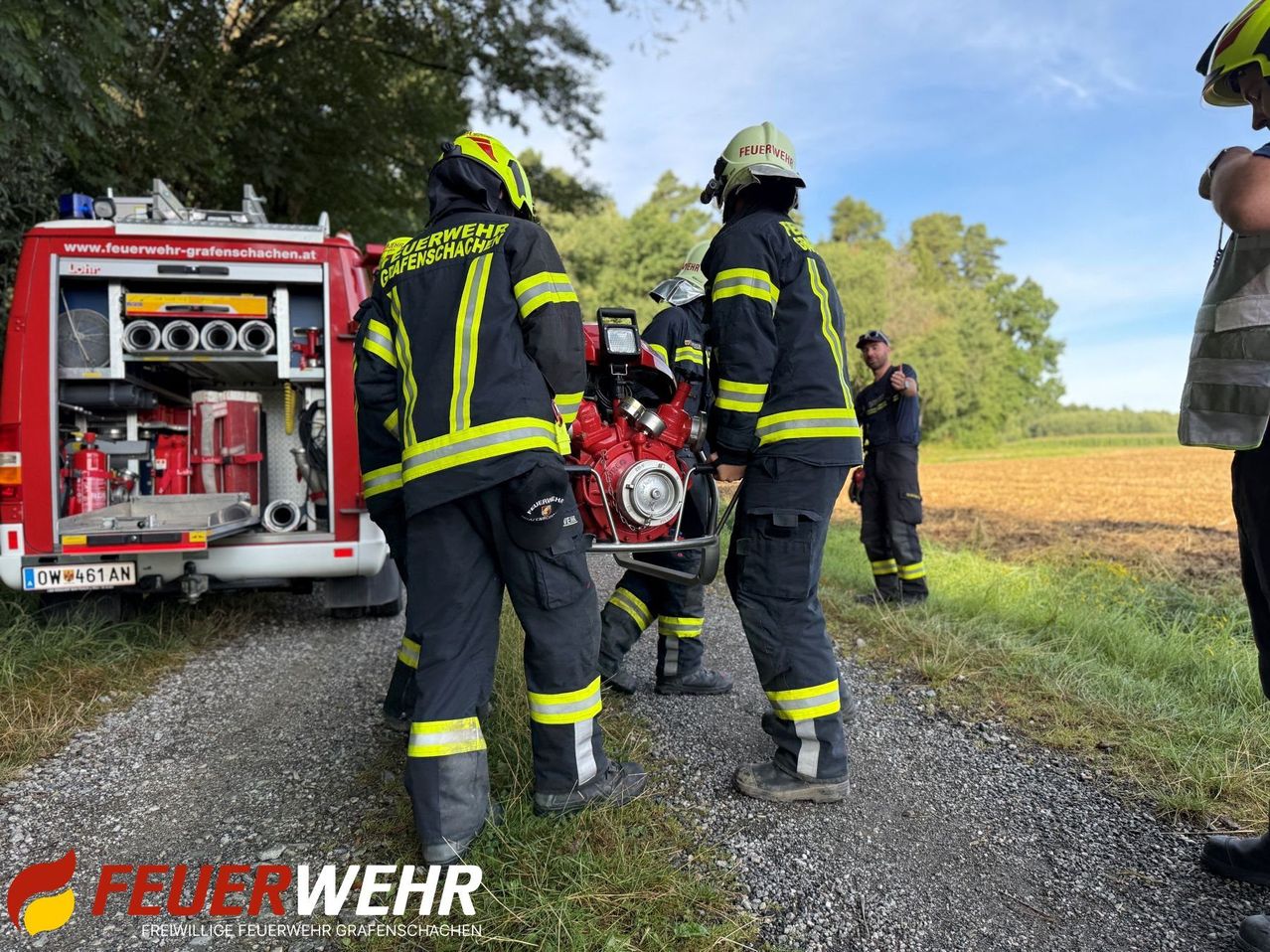 Feuerwehrleute in Uniformen bereiten ein Feuerwehrauto auf einem Kiesweg für den Einsatz vor, während ein anderer Feuerwehrmann beobachtet. Im Hintergrund sind ein Feld und Bäume zu sehen.