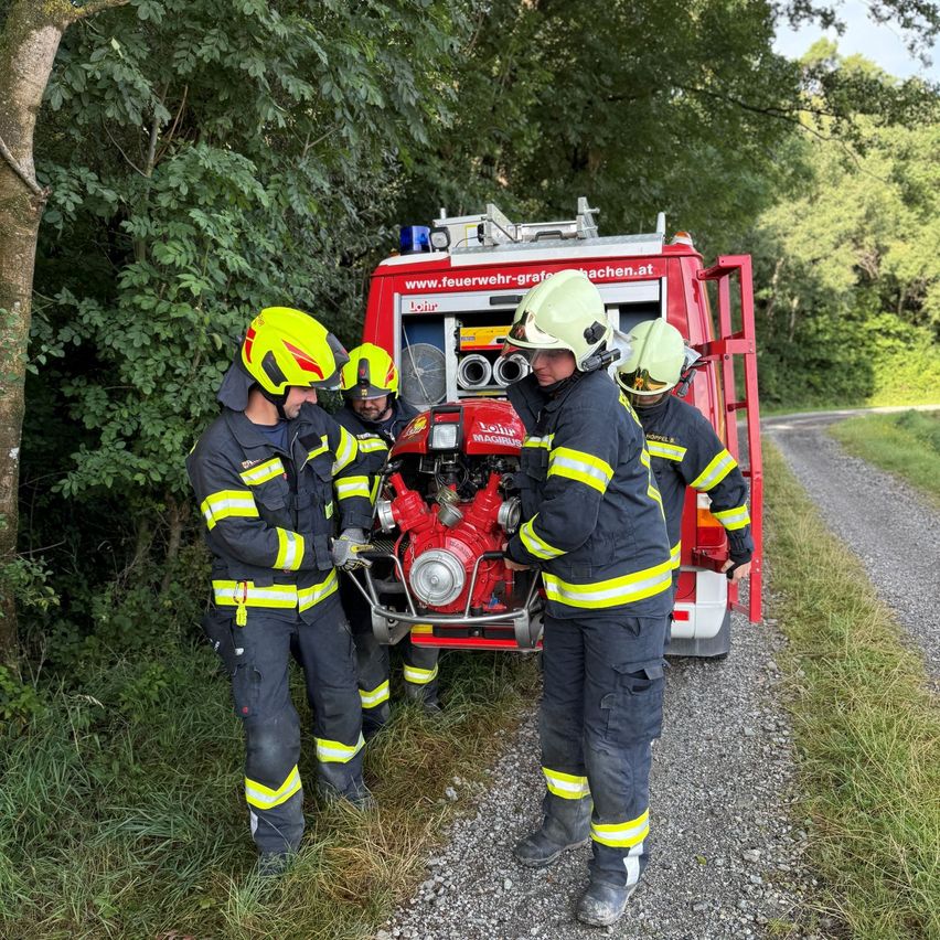 Fünf Feuerwehrleute stehen um einen Feuerwehrwagen auf einer Schotterstraße. Sie tragen Helme und Handschuhe, einer arbeitet an einem roten Gegenstand. Der Wagen ist am Straßenrand geparkt, umgeben von Grün.
