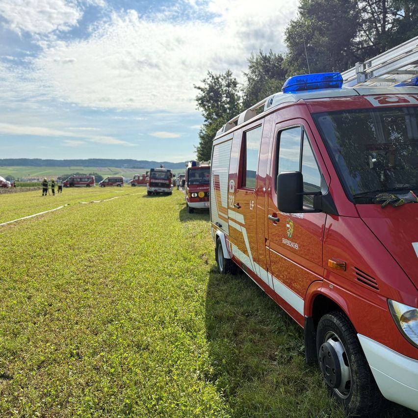 Rettungsfahrzeuge, einschließlich Feuerwehrfahrzeuge, sind auf einem Grasfeld geparkt, mit Menschen im Hintergrund, unter einem teilweise bewölkten Himmel.