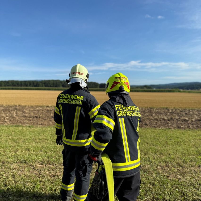 Zwei Feuerwehrleute in voller Ausrüstung stehen auf einem Feld und blicken auf eine landwirtschaftliche Landschaft. Der Himmel ist klar mit einigen Wolken.