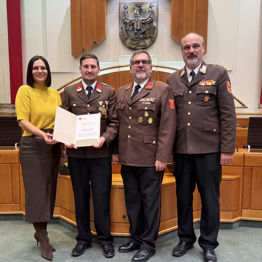 Eine Frau und zwei Männer in Militäruniformen stehen in einem Konferenzraum und halten ein Zertifikat. Hinter ihnen sitzt ein Mann an einem Podium.