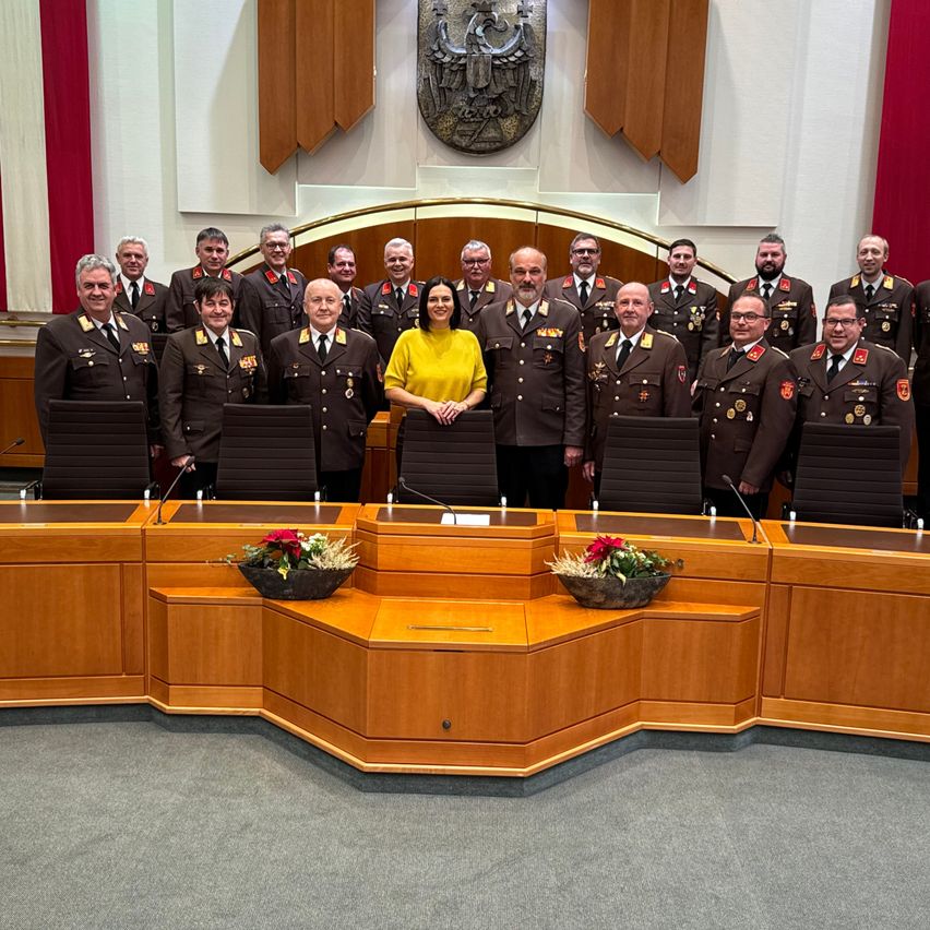 Eine Gruppe uniformierter Personen steht hinter einem langen Holztisch, wahrscheinlich in einem Ratssaal. Eine Frau in einem gelben Oberteil steht in der Mitte. Blumenvasen stehen auf dem Tisch. Dahinter befindet sich eine Wand mit einem dekorativen Bogen und einer montierten Plakette.